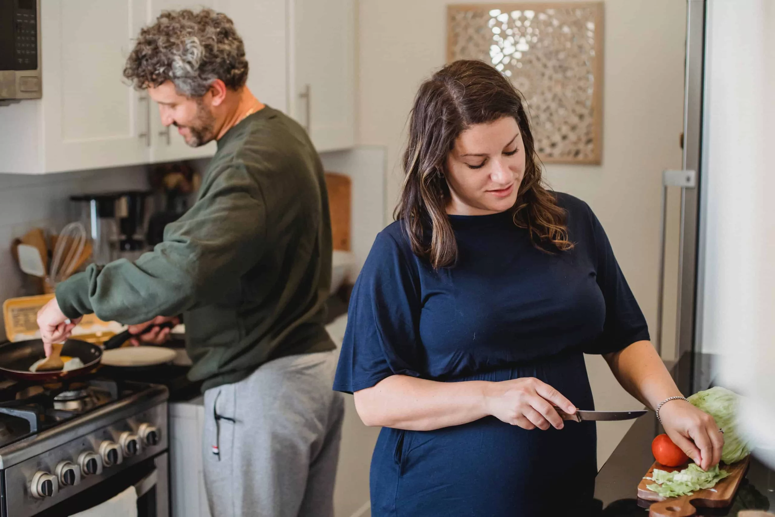 pexels-amina-filkins-5427305 A woman with long brown hair facing cutting something on a chopping board and a man with curly here standing by the cooker and cooking
