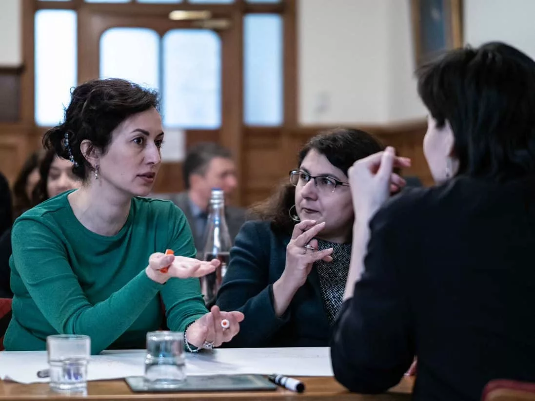 John Smith Trust Fellows from Kazakhstan and Armenia at a discussion. Three women at a table, one of them wearing green jumper, others wearing black. The woman in green jumper is talking and the other two are listening.