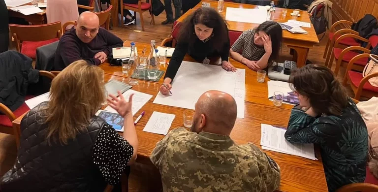 People sitting around a desk during a discussion with two people facing and three people with their backs to the viewer
