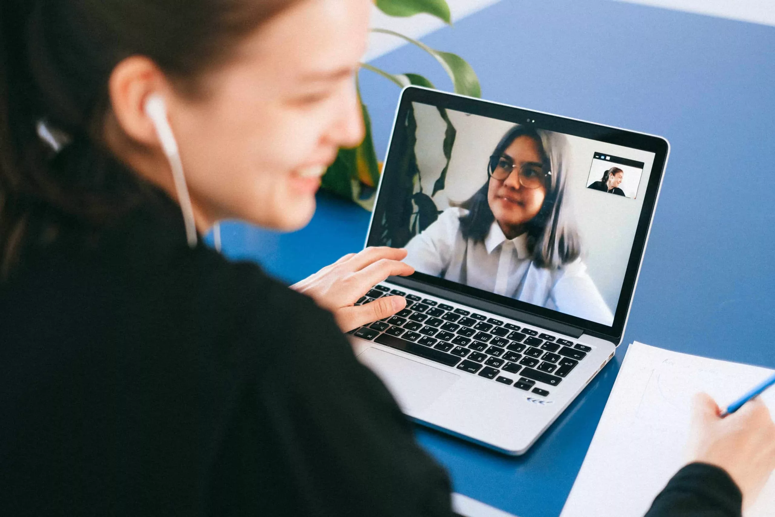 pexels-anna-shvets-4226262 A woman sitting in front of a notebook, with a headphone in her ear, her head turned to us, another woman on the screen of the notebook smiling, they are on a video call