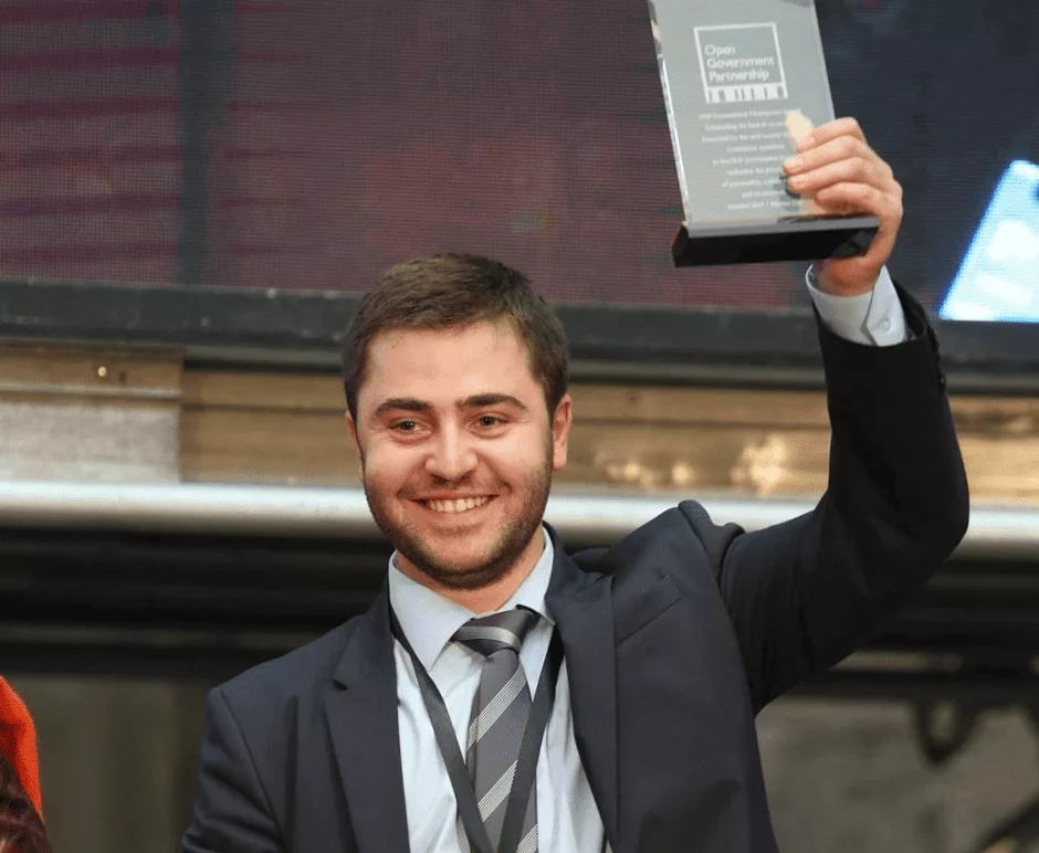 A young man wearing a suit, smiling, holds up a book in his left hand