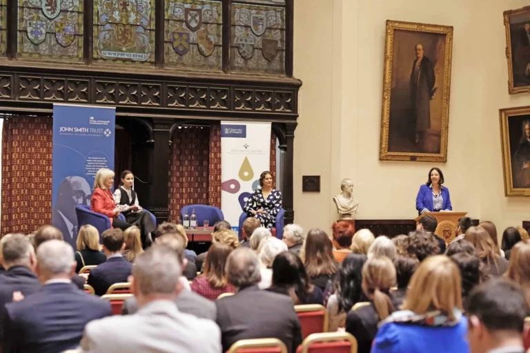A large room full of audience, a panel of speakers at the top