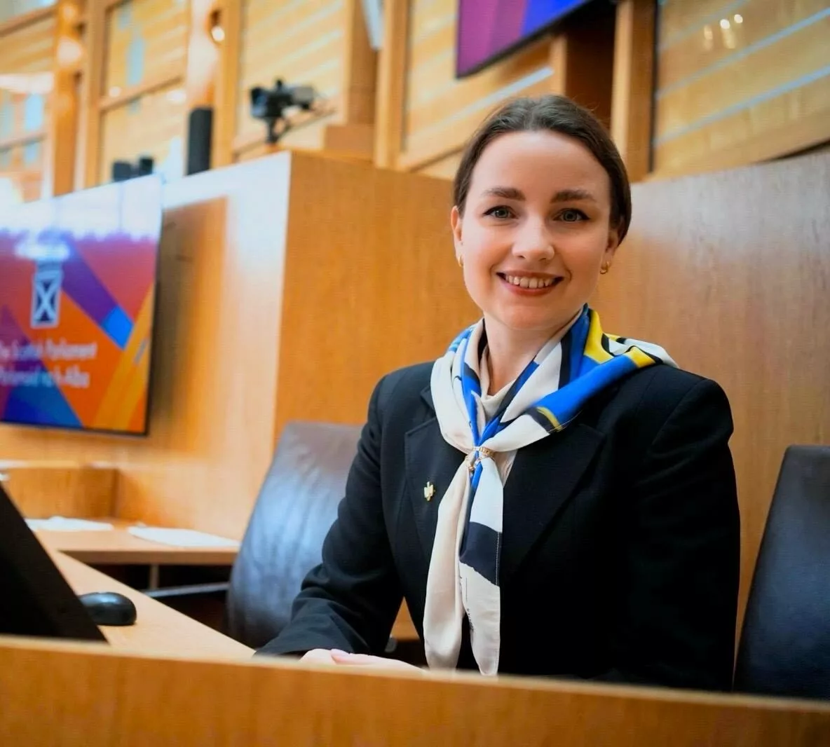 Kateryna Musiienko A woman wearing a dark jacket, sitting at a desk, smiling, with a monitor in front of her