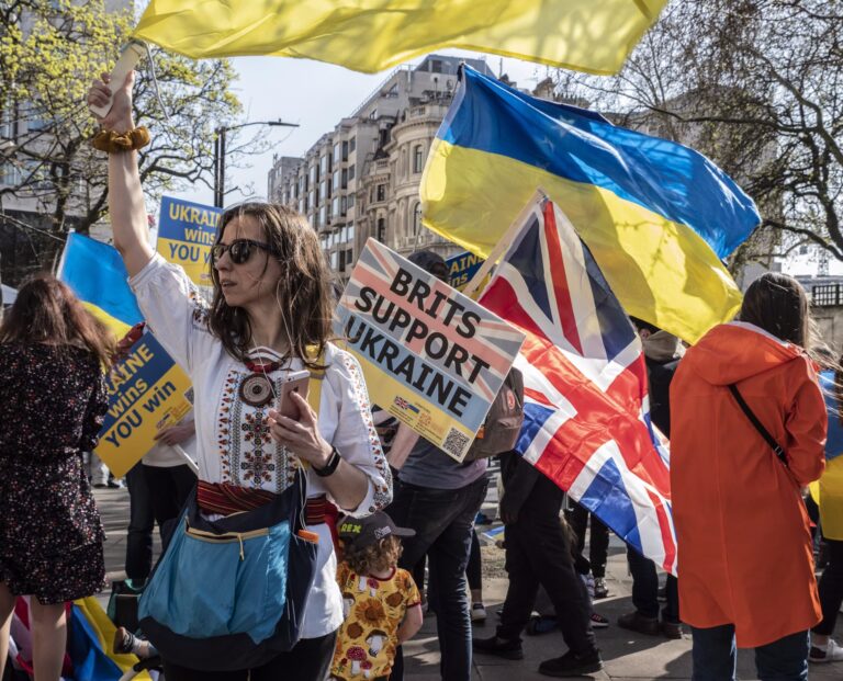 female at a rally raising arm standing in front of British and Ukrainan flags and a sign reading 'Brits support Ukraine'