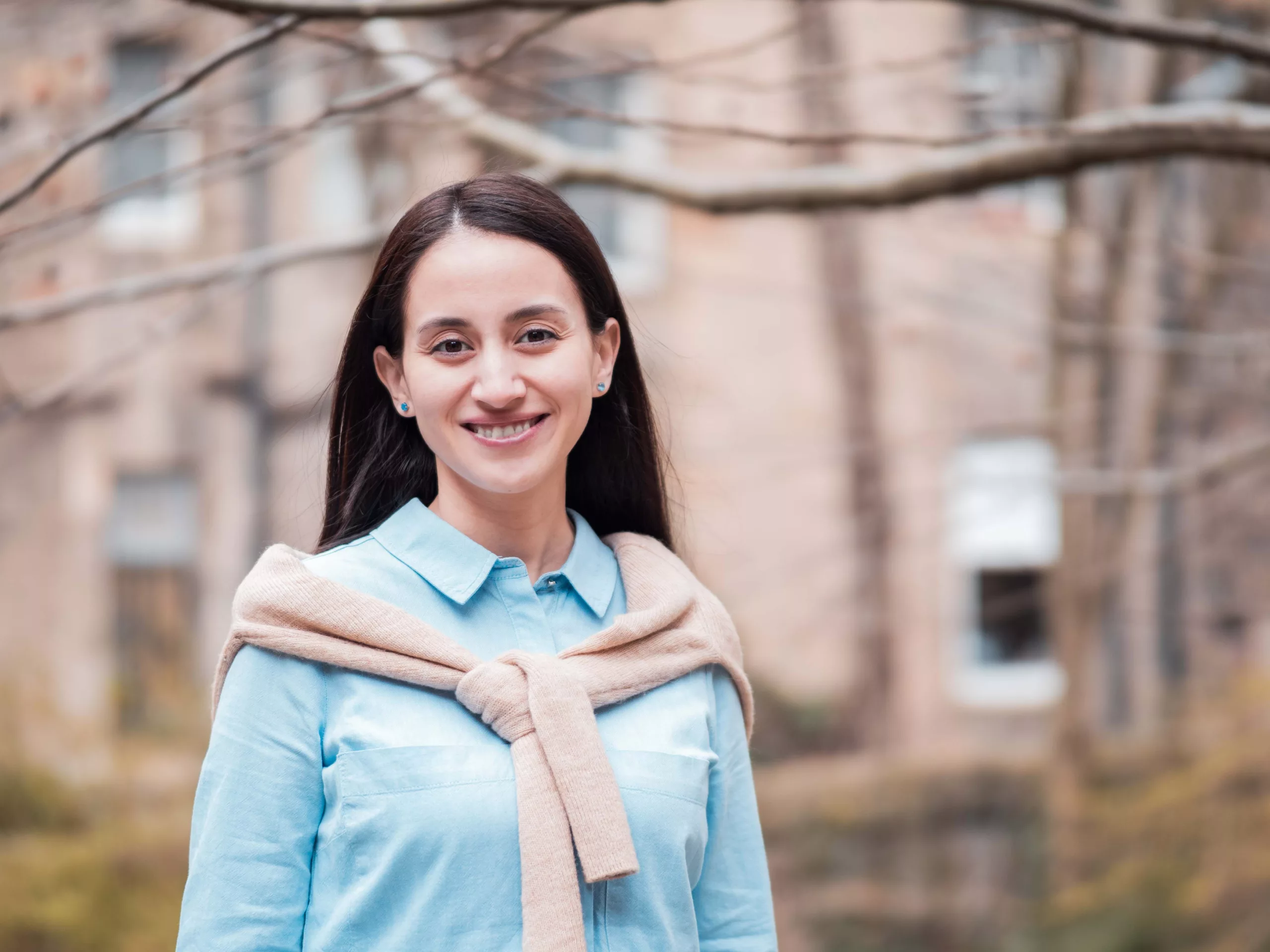 Woman with long dark hair, smiling and standing in front of tree and building