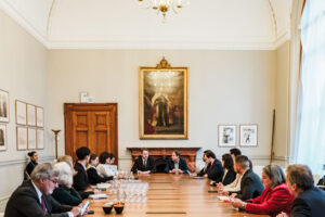 A group of people sat around a large table in a grand room with a painting on the wall