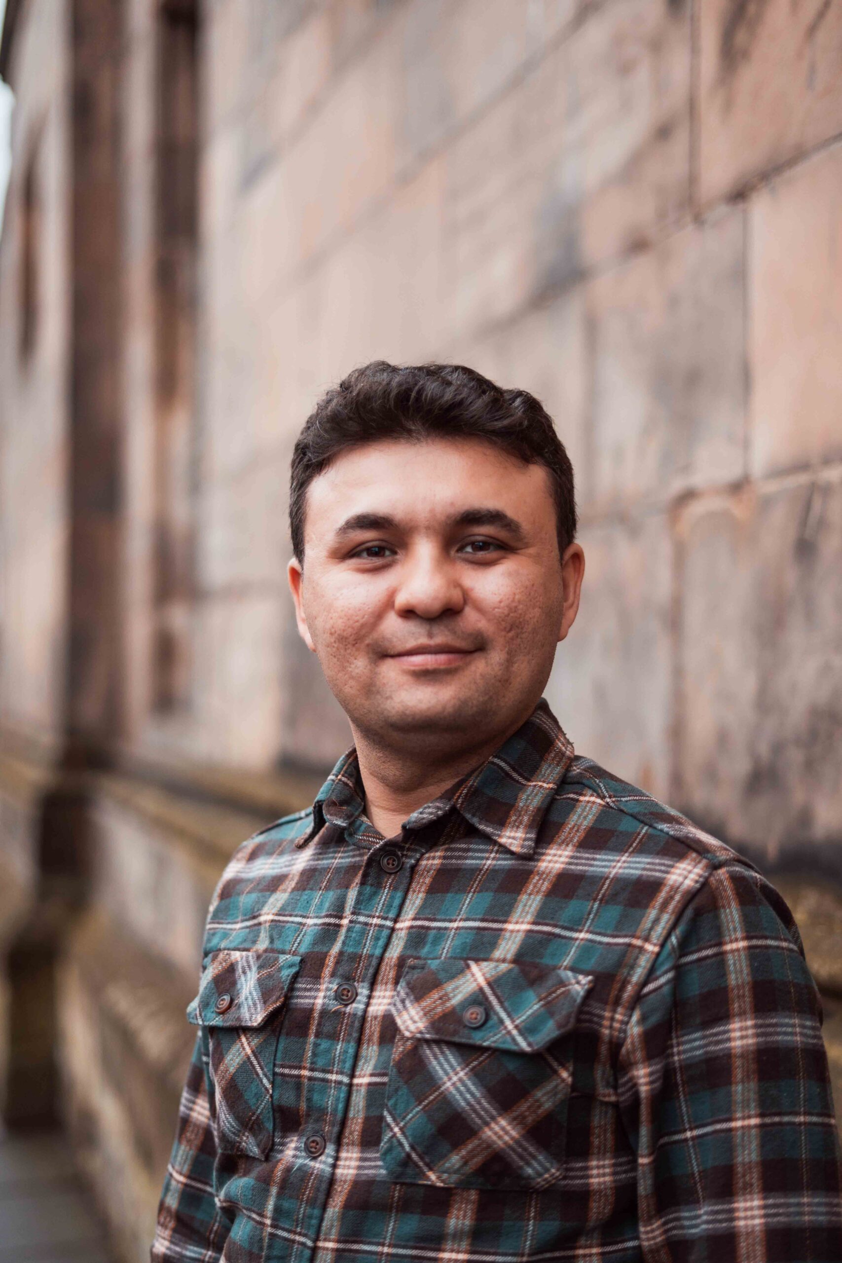 A man in a plaid shirt standing in front of a stone brick building