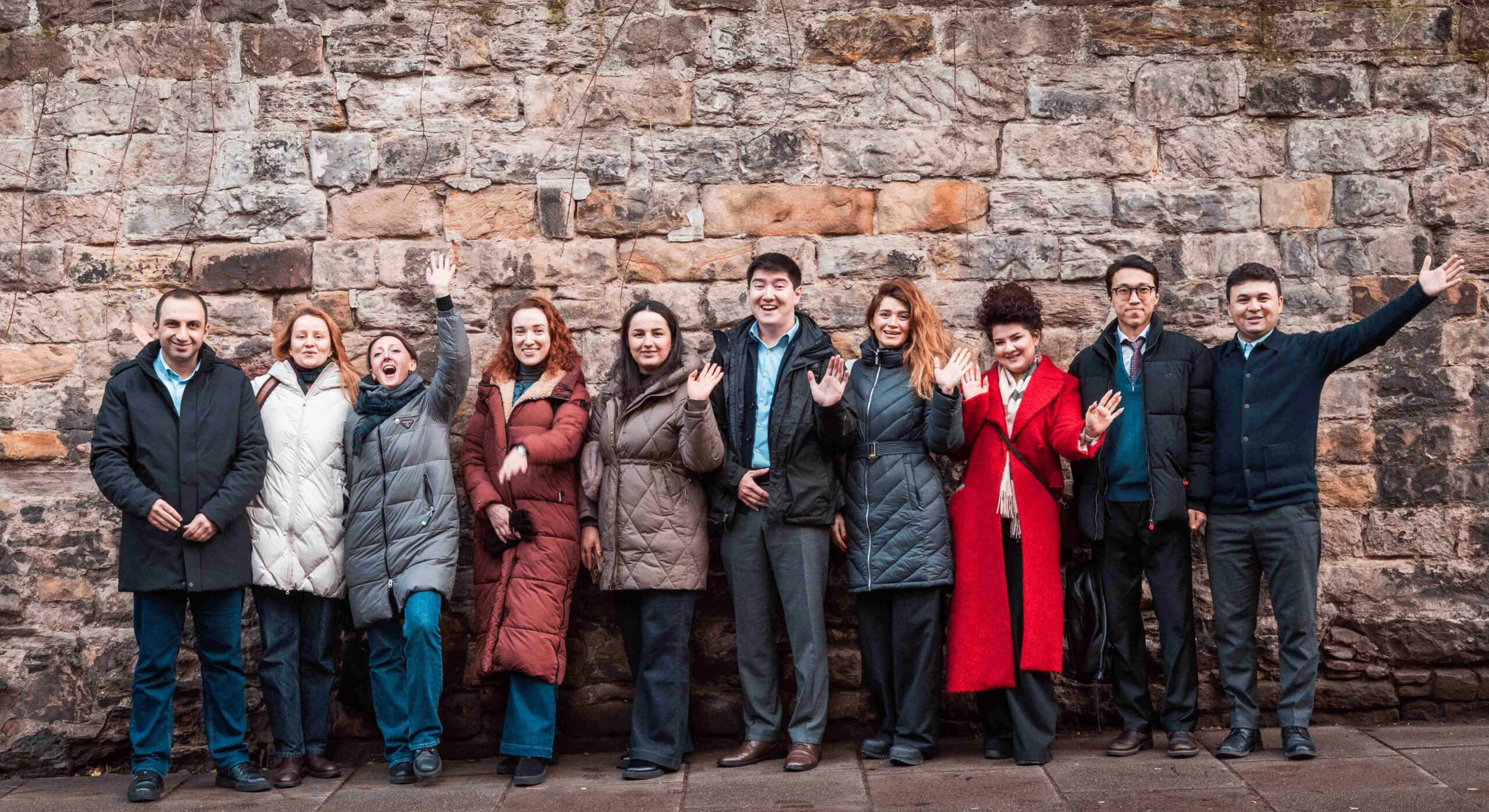 A group of Fellows lined up in front of a brick wall and waving