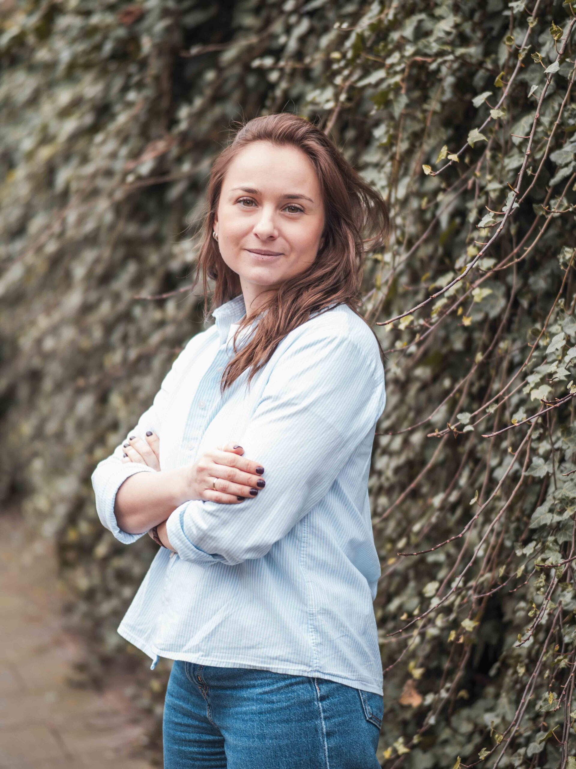 Anna Yafremenka A woman in light shirt and jeans with folded arms standing in front of a wall covered with ivy