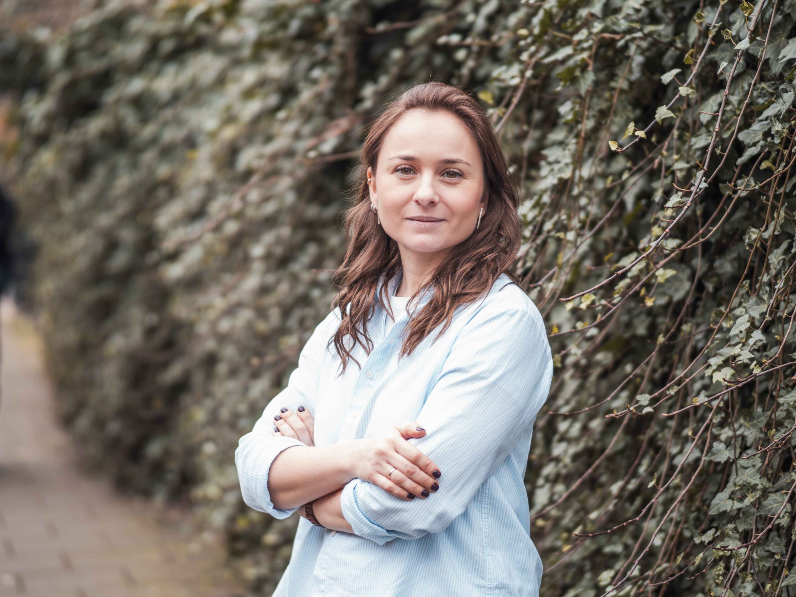 Young woman standing with folded arms in front of a wall of ivy